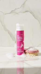 Pink bottle labeled 'HERA CHAMBER' on a marble surface with a brush and pearls.