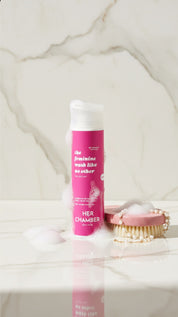 Pink bottle labeled 'HERA CHAMBER' on a marble surface with a brush and pearls.